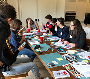 Brill sits at a long table in a common room with six students sitting next to her. She has her hand over her forehead smiling at a student who is gesticulating as he talks.