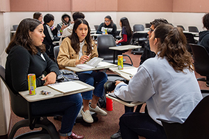 Three female students and one male student sit in a circle with their desks in a classroom with a voter registration drive booklet